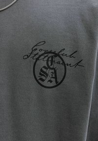 Tissu gris avec une écriture stylisée noire et un logo circulaire, présentant les lettres entrelacées "S" et "A" au centre.