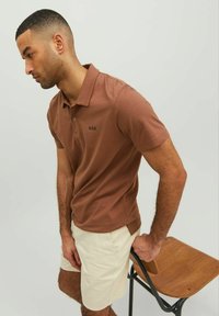 Hnedý polo tričko s jemnou textúrou a zapínacím golierom, s čiernym vyšitým textom 'R.D.D.'; kombinované so svetlo béžovými šortkami.