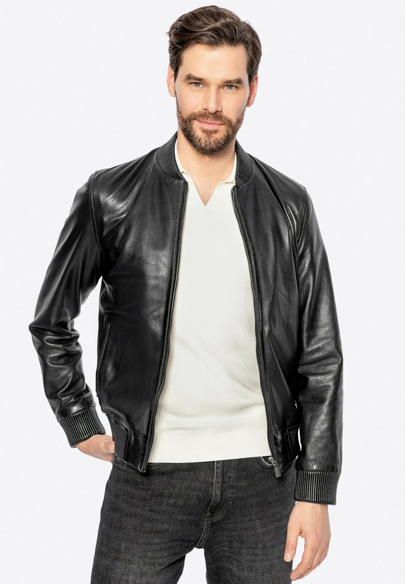 Man met een baard, gekleed in een zwarte leren jas over een wit shirt en donkergekleurde jeans, staand met een hand in zijn zak, neutrale achtergrond.