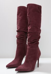 Botas de ante burdeos hasta la rodilla con un diseño arrugado, punteras puntiagudas y tacones de aguja; cuentan con una cremallera lateral para facilitar su uso.