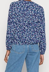 Blommig blus med marin bakgrund, prydd med vita och rosa blommor, långa puffärmar och en rynkad bakdesign. Matchas med ljusblå jeans.