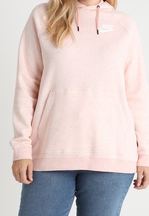 Kvinde iført lyserød Nike-hættetrøje med front-kængurulomme og blå jeans, stående mod en ensfarvet baggrund.