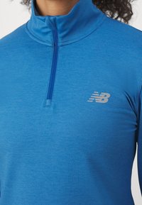 Dlouhý rukáv, modrý top s zipem u krku vyrobený z hladkého, lehkého materiálu, který má malé stříbrné logo na hrudi a vysoký límec.