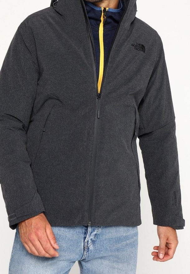 Homme portant une veste zippée gris foncé avec le logo The North Face, un jean bleu clair et une veste marine avec une fermeture éclair jaune en dessous.