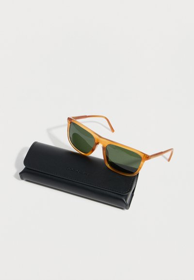 Καφέ διάφανα γυαλιά ηλίου με σκούρους πράσινους φακούς, τοποθετημένα πάνω σε κλειστή μαύρη δερμάτινη θήκη σε λευκό φόντο.