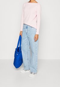 Rosa Langarmshirt, hellblaue hochgeschnittene weite Jeans, weiße Sneakers und eine blaue Tragetasche mit gestreiftem Akzent.