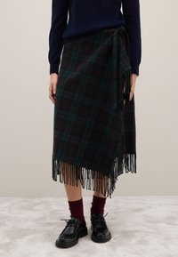 Jupe portefeuille à carreaux verts foncés et noirs avec un ourlet à franges, faite d'un matériau doux. Portée avec un pull bleu marine, des chaussettes bordeaux et des chaussures noires.