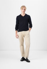 Tengerészkék V nyakú pulóver, kötött anyag; bézs nadrág; fekete cipő; a modell az egyik kezét a zsebében tartja, minimalist fehér háttér előtt.