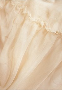 Beige tule stof met een zachte, gestructureerde weving, voorzien van een gerimpelde rand langs de edge. Licht en luchtig uiterlijk met een doorschijnende kwaliteit.
