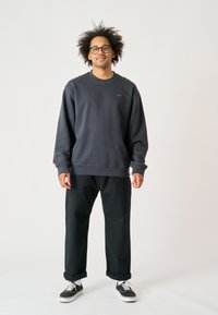 Kohlegrauer Pullover mit gerippten Bündchen und Kragen, ausgestattet mit einem kleinen türkisfarbenen Logo. Kombiniert mit schwarzen Hosen und kontrastierenden Sneakers.