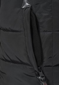 Primo piano di una giacca isolante nera, che mostra una tasca con zip foderata in pile e un esterno in nylon liscio, evidenziato da un bordo della zip rinforzato.