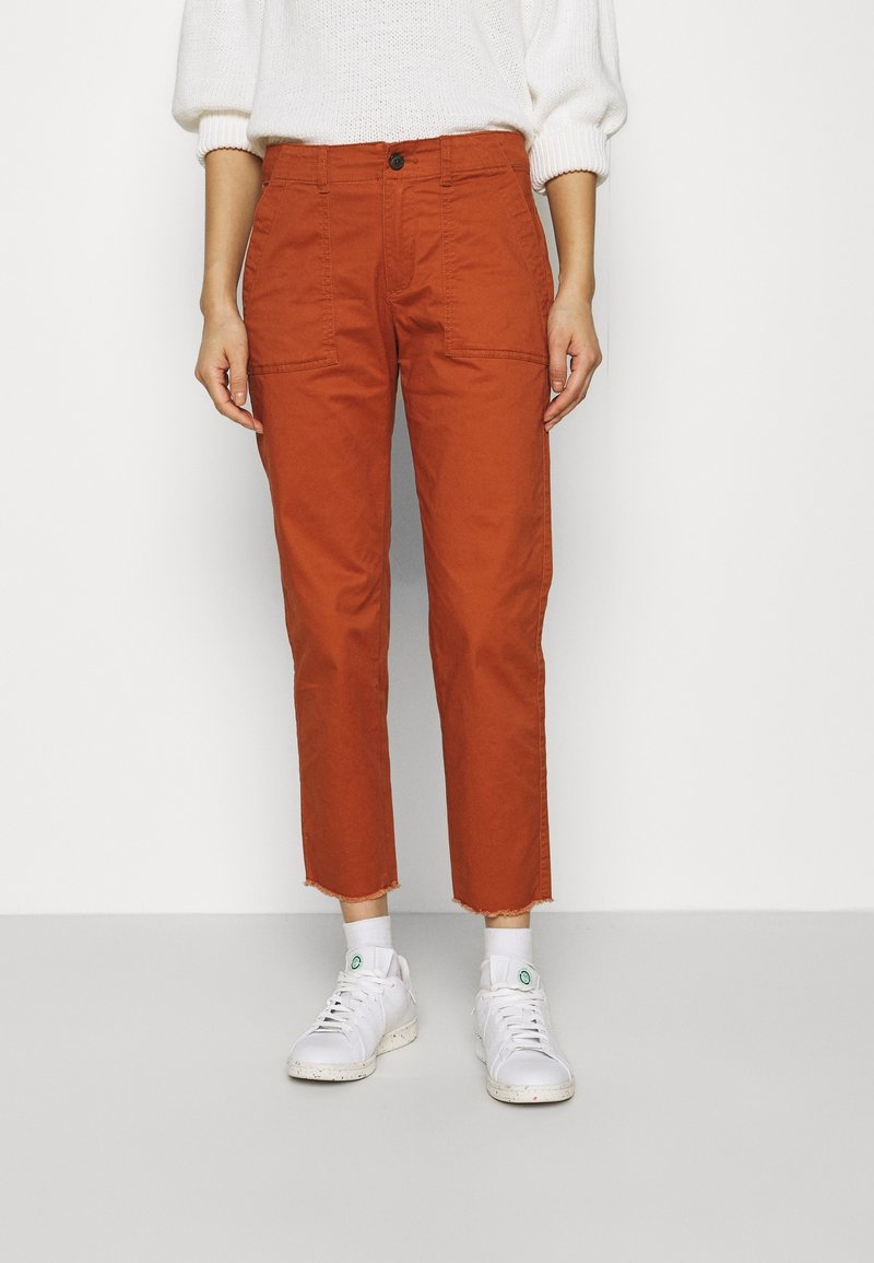 Pantalon couleur rouille, coupe ajustée, avec deux poches avant et un ourlet brut à la cheville. Porté avec des baskets blanches.