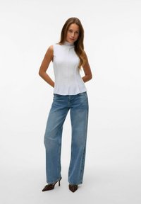 Witte mouwloze top met een hoge hals en textuur, gecombineerd met lichtblauwe wijde jeans en puntige bruine hakken met accenten.