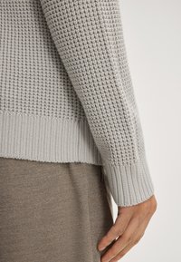 Hellgrauer Strickpullover mit strukturiertem Muster, gerippten Bündchen und Saum. Der Stoff wirkt weich und wird mit einem taupefarbenen Kleidungsstück darunter kombiniert.