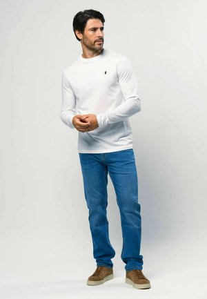Bílé tričko s dlouhým rukávem a kulatým výstřihem s malým černým logem, kombinované s modrými džínami a hnědými teniskami.