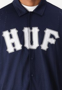 Navyblauw mesh honkbalshirt met grote witte geborduurde "HUF" letters, knoopsluiting en een klassiek kraagontwerp.
