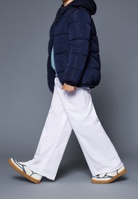 Marineblauwe pufferjas, losse witte broek met wijd uitlopende pijpen, en sneakers met patroon en een gumzool. Model loopt tegen een grijze achtergrond.