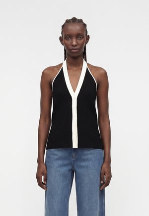 Žena s vrkočmi má na sebe čierne tielko s bielym lemovaním s gombíkmi a modré rifle s vysokým pásom, stojaca na jednotnom pozadí.