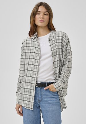 Noor naine, kes kannab lahtist halli ja valget ruudulist nööpidega särki valge t-särgi peal, siniseid teksaseid ja musta vööd.