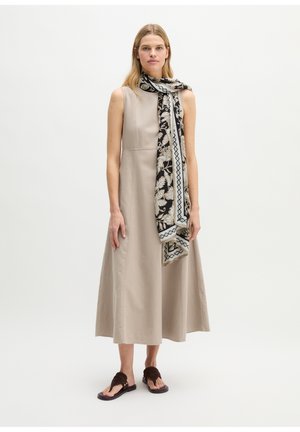 Femme portant une robe midi beige sans manches, une écharpe noire et beige à motifs et des sandales marron foncé, debout devant un fond clair uni.