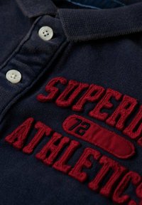 Marineblaues Poloshirt aus strukturiertem Stoff, mit weißen Knöpfen und rot gesticktem Text "SUPER" und "ATHLETIC" sowie Akzentdetails.