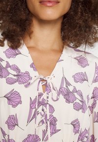 Leichtes Kleid mit einem cremefarbenen Grund und einem lavendelfarbenen Blumenmuster, V-Ausschnitt und einer Schnürung mit passenden Bindebändern.