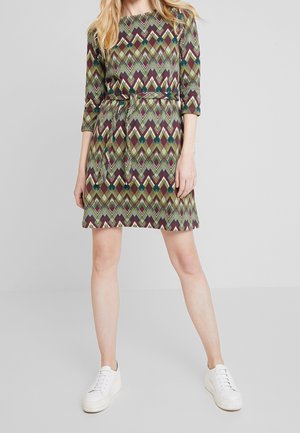 Kobieta w sukience do kolan z geometrycznym wzorem w kolorach zielonym, bordowym i szarym oraz białych trampkach, stoi na tle szarego tła.