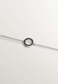 Pulsera de cadena de plata con un colgante circular adornado con piedras negras, creando un acento texturizado en el centro.