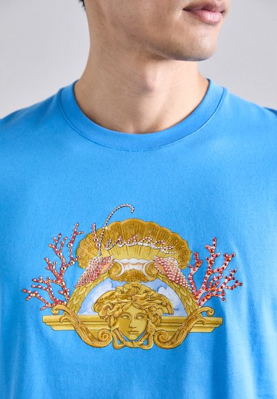 T-shirt en coton bleu avec un design graphique doré et corail, représentant un visage avec des embellissements et le logo "Versace" au-dessus.