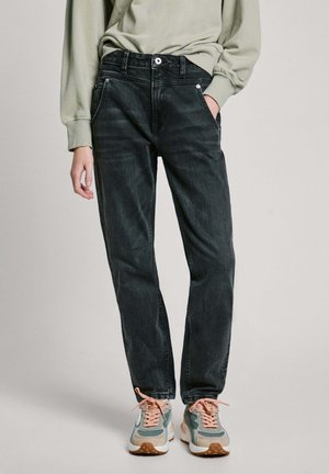 Person trägt dunkel gewaschene Jeans mit geradem Bein, hellgrünes Sweatshirt und mehrfarbige Sneakers mit pfirsichfarbenen Schnürsenkeln, steht vor einem schlichten Hintergrund.