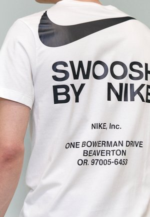 Henkilö pukeutunut valkoiseen Nike-paitaan, jossa on suuri musta swoosh-logo ja teksti "SWOOSH BY NIKE" sekä Niken osoite paidan takana.