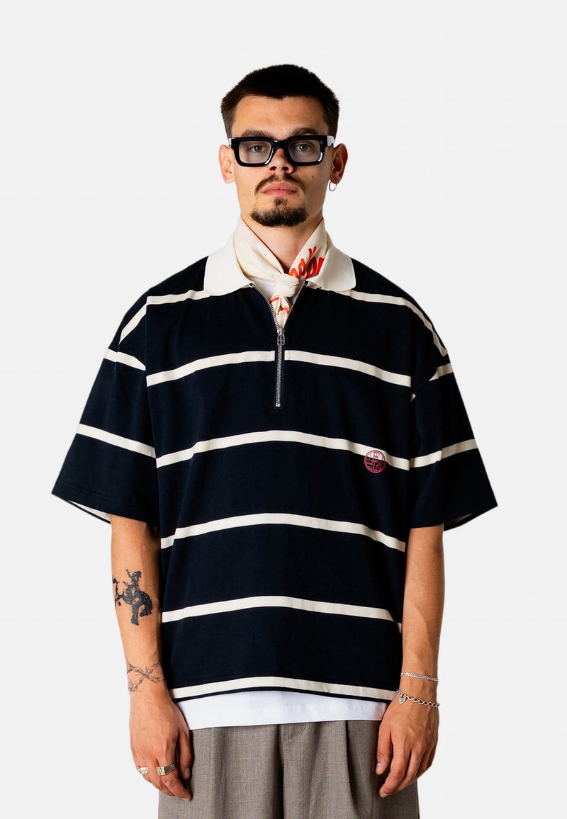 Homme portant un polo à rayures noires et blanches, une écharpe à motifs autour du cou, des lunettes et un pantalon gris, debout devant un fond uni.