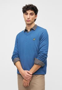 Kék pulóver V-nyakú kialakítással, texturált szövettel, és egy kis, sárga logóval a mellrészen. Egy galléros szürke inggel van aláöltve.