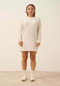 Dzianinowa sukienka w kolorze kremowym z prążkowanymi mankietami i dołem. Posiada teksturowany wzór. Modelka nosi kremowe sneakersy i białe skarpetki do kostek.
