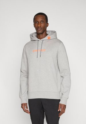 Mann trägt hellgrauen Hoodie mit orangefarbenem "FIRE+ICE" Schriftzug auf der Brust und schwarze Hose, steht vor einfarbig hellem Hintergrund.