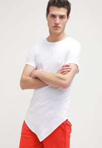 Camiseta blanca de algodón con dobladillo redondeado, mangas cortas y textura suave, combinada con pantalones cortos rojos. El modelo está de pie con los brazos cruzados.
