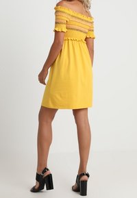 Žluté šaty bez ramínek s nařaseným korzetem a volány na okrajích, se rovně střiženým lemem a doplněné černými sandály na vysokém podpatku.