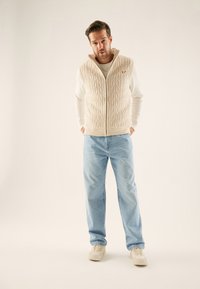 Gilet en tricot beige avec un motif en câble, fermeture éclair, porté sur une chemise blanche à manches longues, assorti à un jean bleu clair et des baskets beiges.