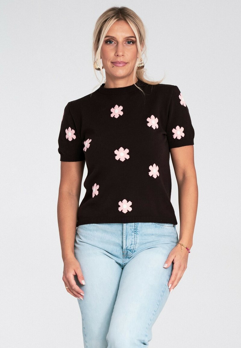 Blondynka nosząca złote kolczyki w kształcie kół, czarny sweter z krótkim rękawem z różowymi kwiatami oraz jasnoniebieskie dżinsy na jednolitym tle.