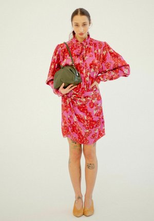 Abito floreale in tonalità rosa e rosso con maniche a palloncino lunghe, dettaglio annodato in vita, abbinato a una borsa verde scuro e scarpe beige.