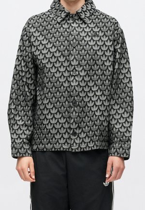 Person trägt eine schwarz-grau gemusterte, geknöpfte Jacke mit wiederholten Logodesigns und schwarze Hose mit kleinem Logo am linken Oberschenkel.