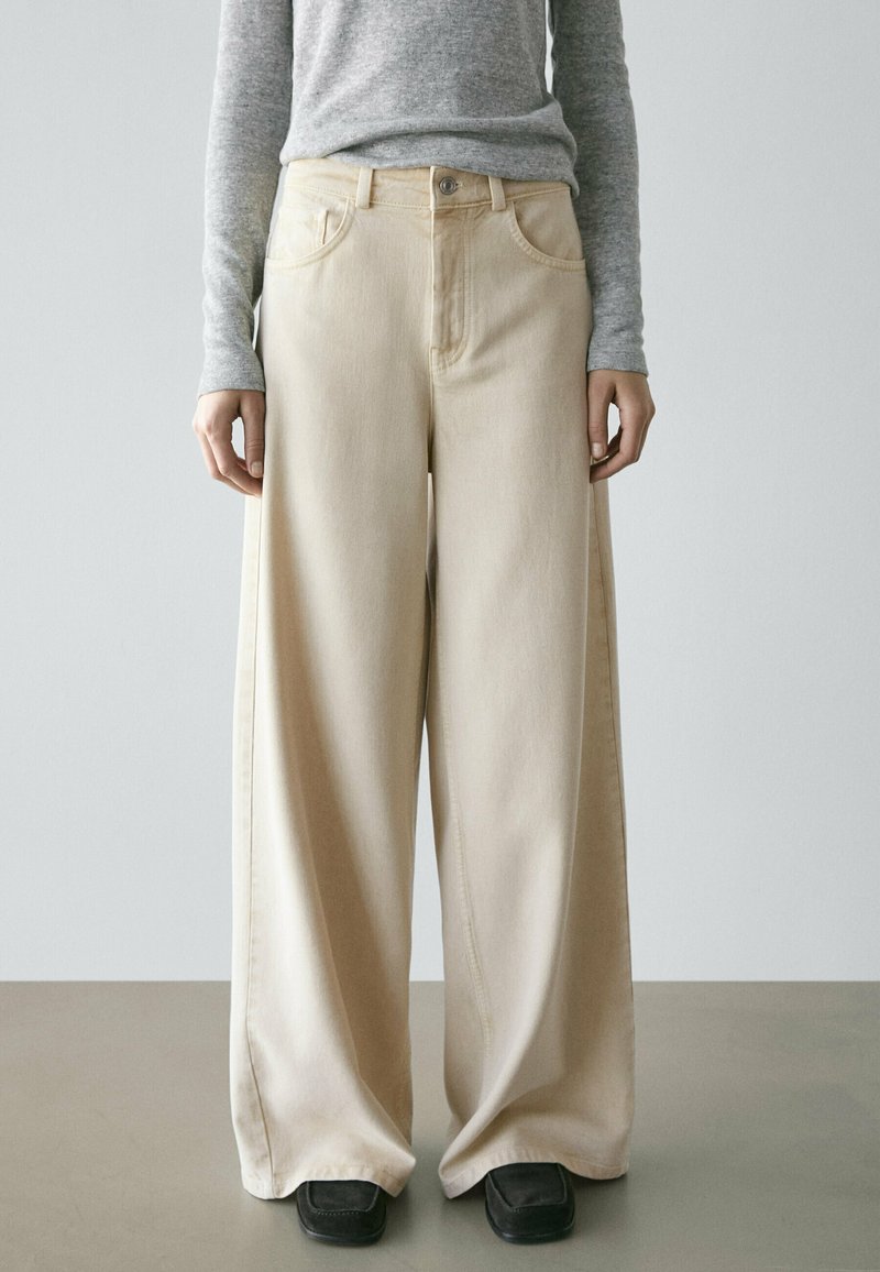 Person trägt beige, weit geschnittene Hose, grauen Langarmshirt und schwarze Schuhe mit quadratischer Spitze und steht vor einem einfarbigen Hintergrund.