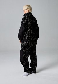 Zwarte oversized jas en broek, met een golvend wit patroon. De stof heeft een gladde textuur, met een hoge kraag en een ontspannen pasvorm.