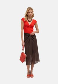 Haut rouge à côtes avec un décolleté en V, associé à une jupe portefeuille marron, portant un sac à main rouge. Porté avec des sandales à talons orange.