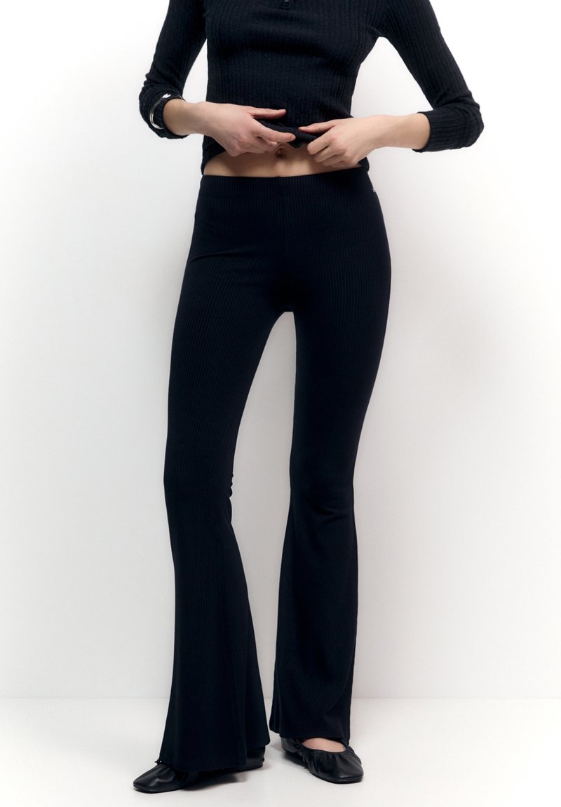 Personne portant un pantalon évasé côtelé noir, un haut noir à manches longues et des ballerines noires, ajustant son haut pour dévoiler le ventre.