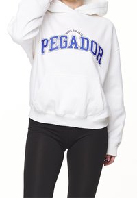 Sudadera blanca con un gran gráfico de texto azul "PEGADOR", con un bolsillo tipo canguro y puños acanalados, combinada con leggings negros.