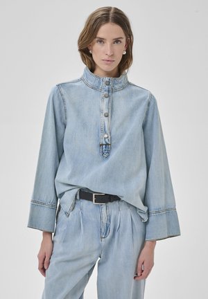 Ung kvinde iført lyseblå denimskjorte med høj krave og knapper, kombineret med matchende højtaljede denimbukser og sort bælte.