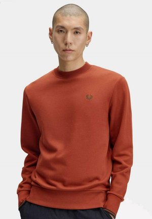 Hombre con cabello corto y pendientes, que lleva una sudadera de color óxido con un pequeño logo de corona de laurel en el pecho.