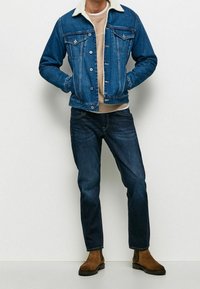 Jeansjacka med fårskinnskrage, blå, knappstängning, två bröstfickor, stylad över en ljus tröja och kombinerad med mörka jeans och bruna stövlar.