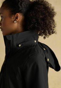 Jachetă impermeabilă neagră cu un guler înalt și nasturi aurii. Caracteristicile includ o glugă și un material texturat, prezentată din lateral.